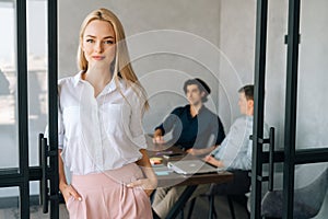 Portrait of attractive blonde female office worker in casual clothes standing in office room, confident looking at