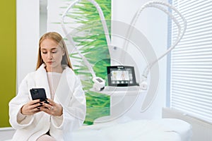 Portrait of attractive blonde female patient in white bathrobe using typing smartphone in modern health center while