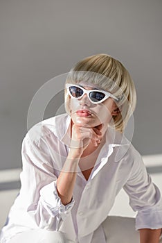 portrait of attractive blonde woman in sunglasses and fashionable white outfit resting chin on hand and looking away
