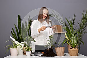 Portrait of attractive brown-haired Caucasian woman botanist working in her orangery and making photo on mobile phone for her blog