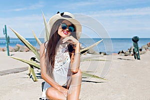 Portrait of attractive brunette girl with long hair sitting on the beach near cactus on the background. She wears hat