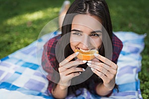 Portrait of attractive brunette woman lying on green grass in outdoor eating orange fruit, copy space for your advertising message
