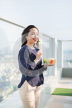 Portrait of attractive caucasian smiling woman eating salad on a sunny summer day sitting on green grass in park, focus on fork