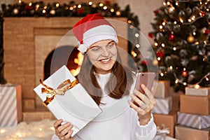 Portrait of attractive Caucasian woman wearing white sweater and santa claus hat, sitting near fireplace and xmas tree with smart