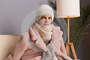 Portrait of attractive Caucasian young adult woman wearing warm clothes and wrapped in scarf sitting on sofa in room, looking at