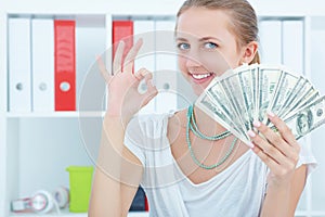 Portrait of attractive cheerful female showing many banknotes of one hundred dollars on the office background.