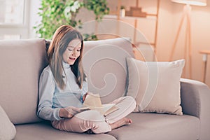 Portrait of attractive cheerful focused preteen girl sitting on divan resting reading book at house flat indoor