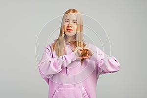 Portrait of attractive cheerful girl showing heart shape sign amour, standing in trendy hoody over gray background. Sign of love,