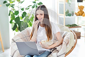 Portrait of attractive cheerful girl sitting in chair watching video eating snack pastime at home light living room