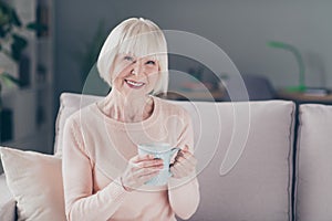 Portrait of attractive cheerful grey-haired woman sitting on divan drinking beverage in house flat indoors