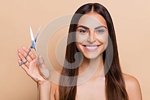 Portrait of attractive cheerful long-haired girl holding in hand scissors isolated over beige pastel color background