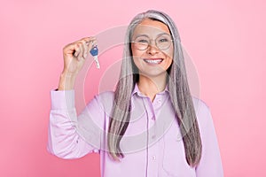 Portrait of attractive cheerful successful gray-haired woman holding house key isolated over pink pastel color