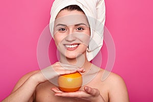 Portrait of attractive cheerful woman with white towel on head, holds orange over pink background. Young smiling female visits spa