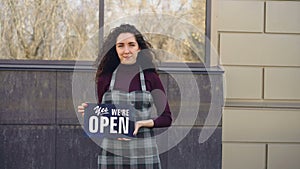 Portrait of attractive confident woman in apron small business owner holding `yes we are open` sign standing outside and