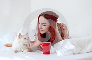 Portrait of an attractive, contented, young, sexy red-haired woman lying relaxed in bed enjoys her coffee in a red cup and cuddles