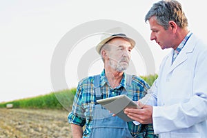 Attractive Crop scientist wearing lab coat showing digital tablet to senior farmer against corn plant growing in field