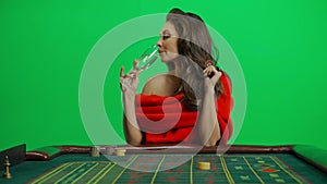 Portrait of attractive female on chroma key green screen close shot. Woman in red dress at roulette table drinks