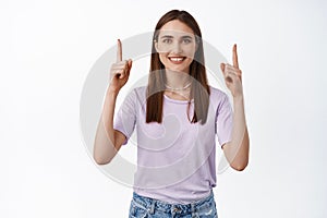 Portrait of attractive female model pointing fingers up, smiling candid, standing in t-shirt, showing advertisement