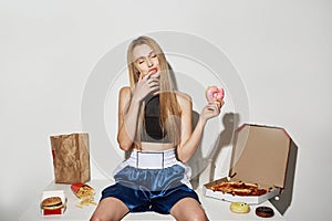 Portrait of attractive girl with blonde hair in sport top and shorts sitting on table with big amount of unhealthy food