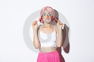 Portrait of attractive girl blowing pink bubble gum on face and looking silly, standing in anime wig and party outfit