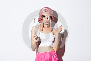 Portrait of attractive girl cosplay fairy for halloween, wearing pink wig and holding magic wand, standing over white