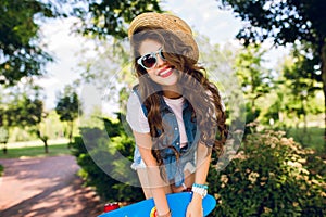 Portrait of attractive girl with long curly hair in hat posing with skateboard in summer park. She wears jeans jerkin