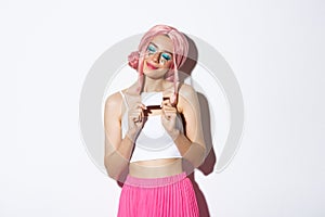 Portrait of attractive girl in pink wig, dreaming about shopping, holding credit card and smiling pleased, standing over