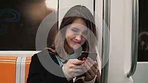 Portrait of attractive girl in subway train using smartphone. Young woman chatting with friends and smiling.