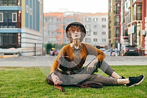 Portrait of attractive girl walking with dog, sitting on lawn, stroking puppy and posing at camera against cityscape background.