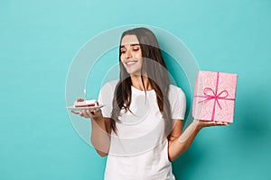 Portrait of attractive happy girl, celebrating birthday and receiving gift, making wish on bday cake, standing over blue
