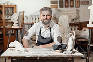Portrait of attractive joyful bearded male sculptor which sitting on the workshop interior background and looks at the