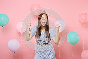 Portrait of attractive joyful young woman in birthday hat blue dress doing winner gesture screming on pink background