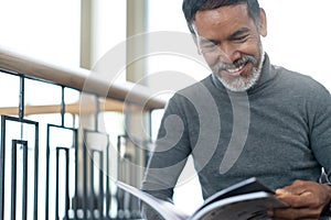 Portrait of attractive mature asian man retired with stylish short beard sitting, smiling and reading magazine books
