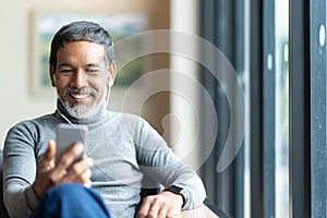 Portrait of attractive mature asian man retired with stylish short beard using smartphone sitting or listening music