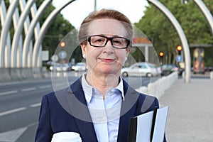 Portrait of attractive mature businesswoman commuter smiling in busy city, holding a coffee cup and paperwork folder