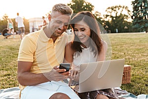 Portrait of attractive middle-aged couple holding cellphone while using laptop computer during picnic in summer park