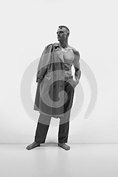 Portrait with attractive muscular man standing and holding coat on shoulder over white studio background. Monochrome