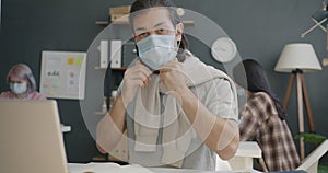 Portrait of attractive office worker putting on medical face mask and looking at camera in workplace