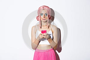 Portrait of attractive party girl in pink wig, with bright makeup, holding smartphone and looking at camera confident