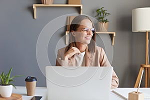 Portrait of attractive positive adorable young businesswoman working online on notebook in home office, looking away with charming