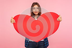 Portrait of attractive positive girl holding big red paper heart and looking at camera with kind smile