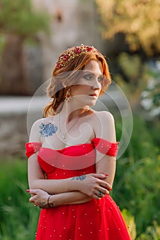 Portrait of attractive redhead tattooed woman in red dress and diadema on blurred medieval castle background