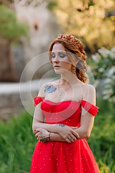 Portrait of attractive redhead tattooed woman in red dress and diadema on blurred medieval castle background