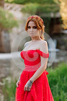 Portrait of attractive redhead tattooed woman in red dress and diadema on blurred medieval castle background