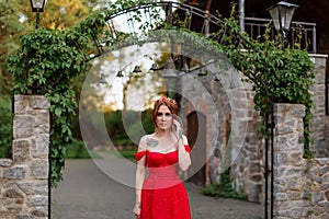 Portrait of attractive redhead tattooed woman in red dress and diadema on blurred medieval castle background