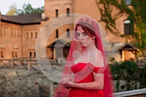 Portrait of attractive redhead tattooed woman in red dress, diadema and red veil on blurred medieval castle background