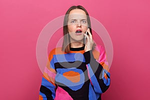 Portrait of attractive scared shocked surprised woman wearing colorful jumper, standing and talking on mobile phone, looking at