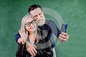 Portrait of attractive senior couple, standing at home in front of green wall and talking with their friends via phone