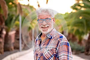 Portrait of attractive senior man white-hair and beard looking at camera. Standing outdoor in a public park at sunset. A smiling