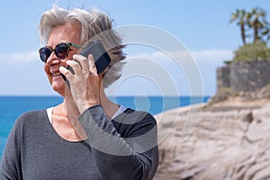 Portrait of attractive senior woman white-haired using smartphone outdoors at the sea smiling happy. Joyful lifestyles concept,